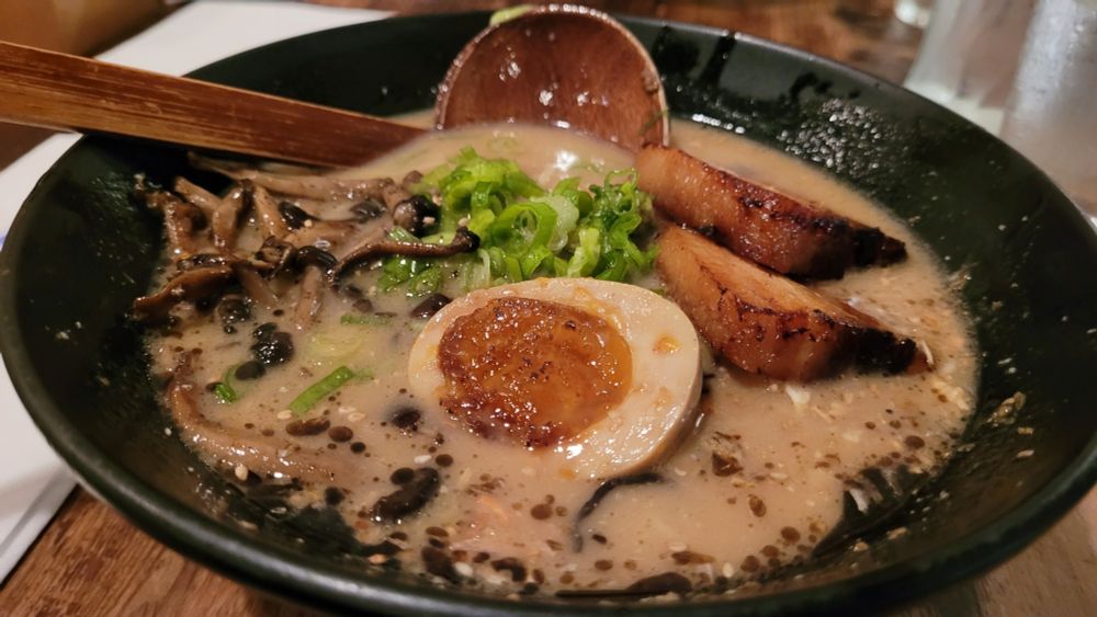 A bowl of ramen with a creamy beef stock, cooked egg, chopped mushrooms, diced scallops, and 2 slices of smoked pork belly. A decently sized wooden ladle is placed inside the bowl to help you eat.
