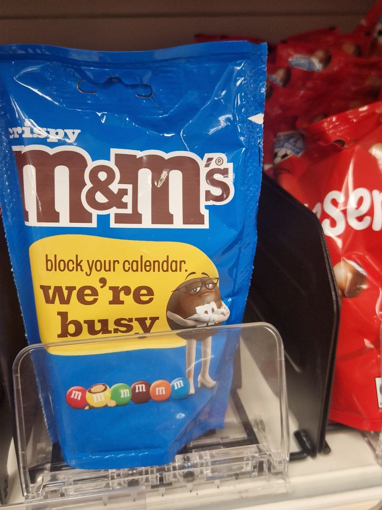 front of a packet of Crispy M&M's the Brown M&M (a humanoid candy wearing high heels and glasses) is saying"block your calendar. We're busy"