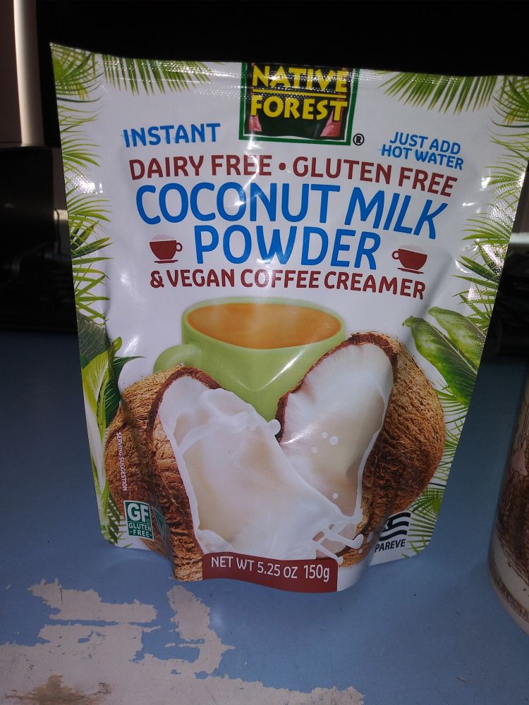 A pouch of Native Forest coconut milk powder. Label shows a halved coconut, coconut milk splashing in the foreground, with a green coffee cup in the background. Blue and brown lettering says "instant. Just add hot water. Dairy free Gluten free Coconut Milk Powder & Vegan coffee creamer."