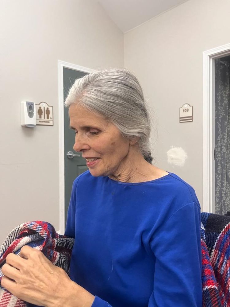 Another photo of my mom.  Her braid is visible at her neck because she's looking forward and is turned a bit compared to the previous photo. She wears the same bright blue fleece long sleeve shirt, and is seated on the same blue and red plaid blanket. She looks at something in her hands which are also holding the plaid. Her eyes and her mouth are open.