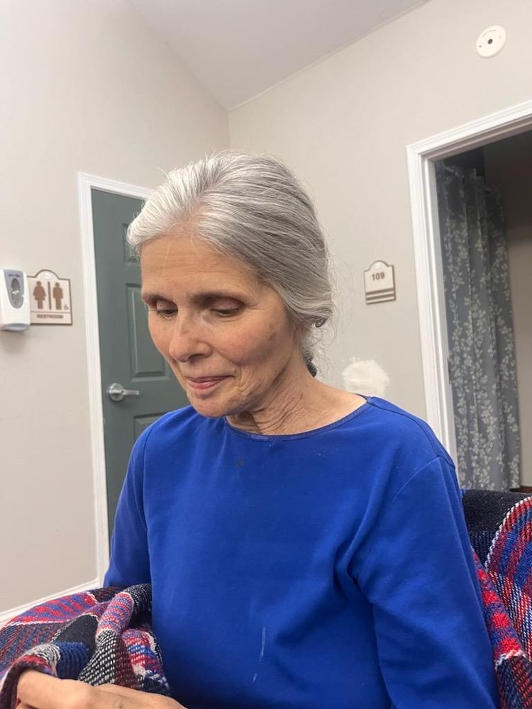 Photo of my mom a 70 year old woman with gorgeous white hair braided down her back. The braid isn't visible. She wears a bright blue fleece long sleeve shirt, and is seated on blue and red plaid blanket. She looks down at something in her lap, with her mouth closed.