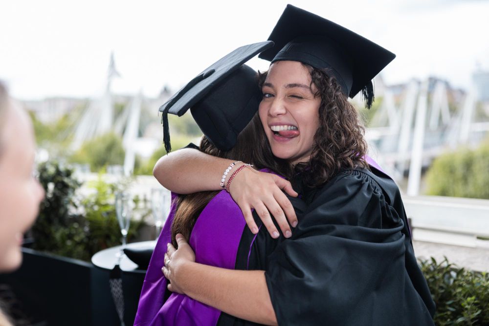 Graduate in cap and gown joyfully hugging another person at an outdoor graduation ceremony.