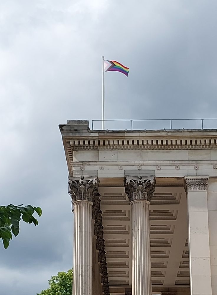 A Pride flag flies atop a classical building with tall columns and cloudy sky in the background