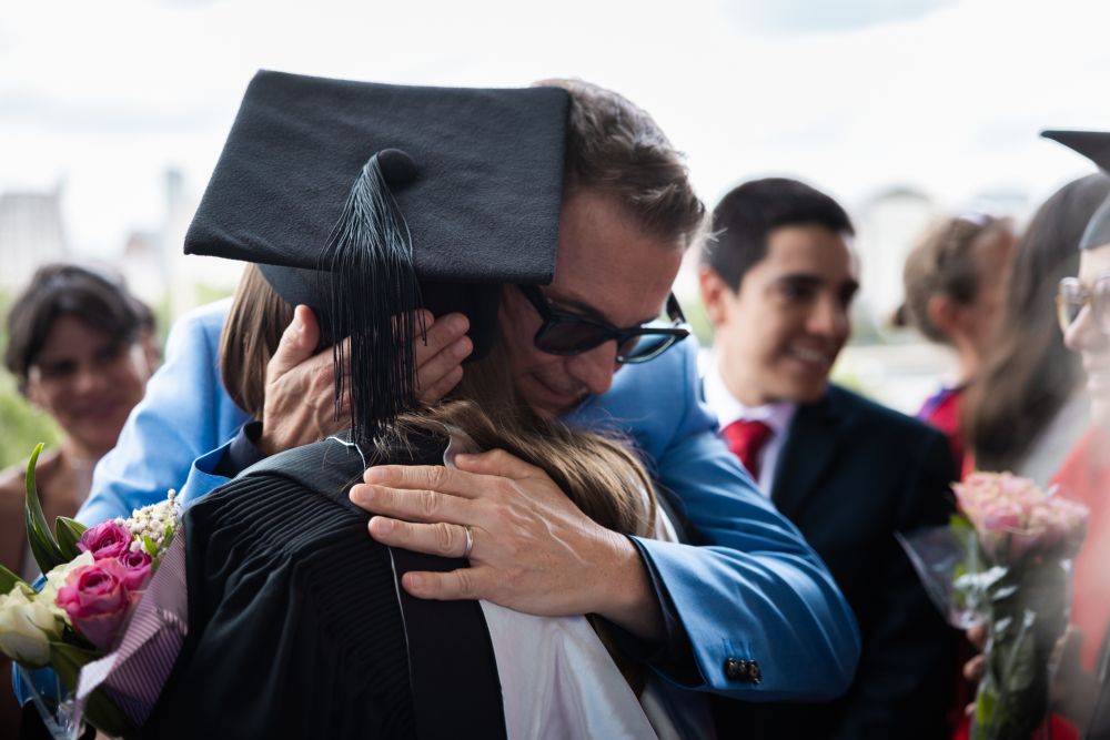 Graduate in a cap and gown embracing another person at a graduation ceremony, with other attendees in the background.