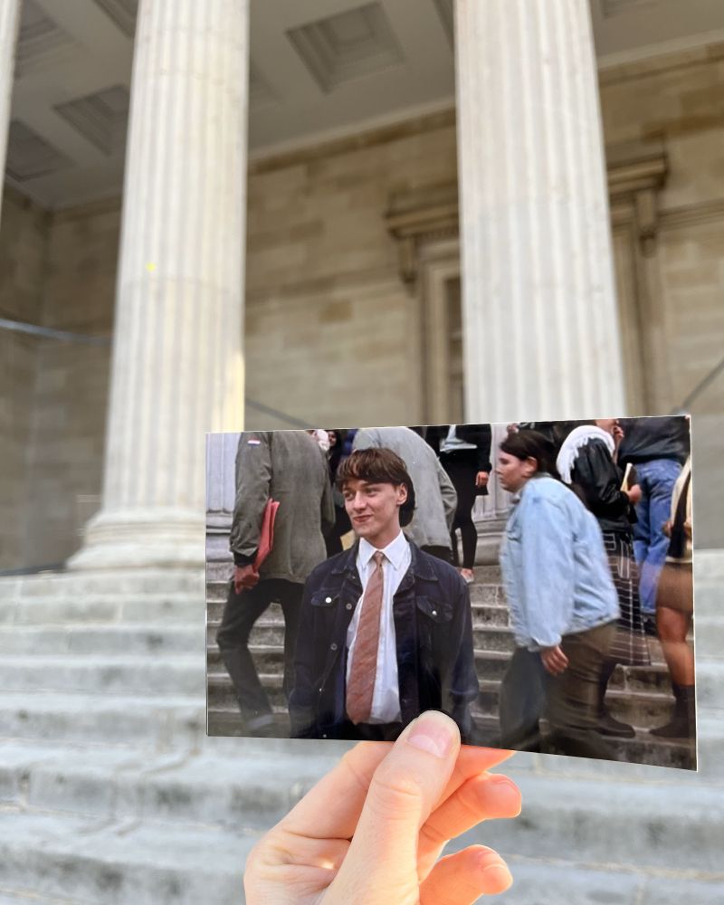 A person holds up a photograph of a scene from the movie "Starter of 10",  aligning it with the actual filming location visible in the background.