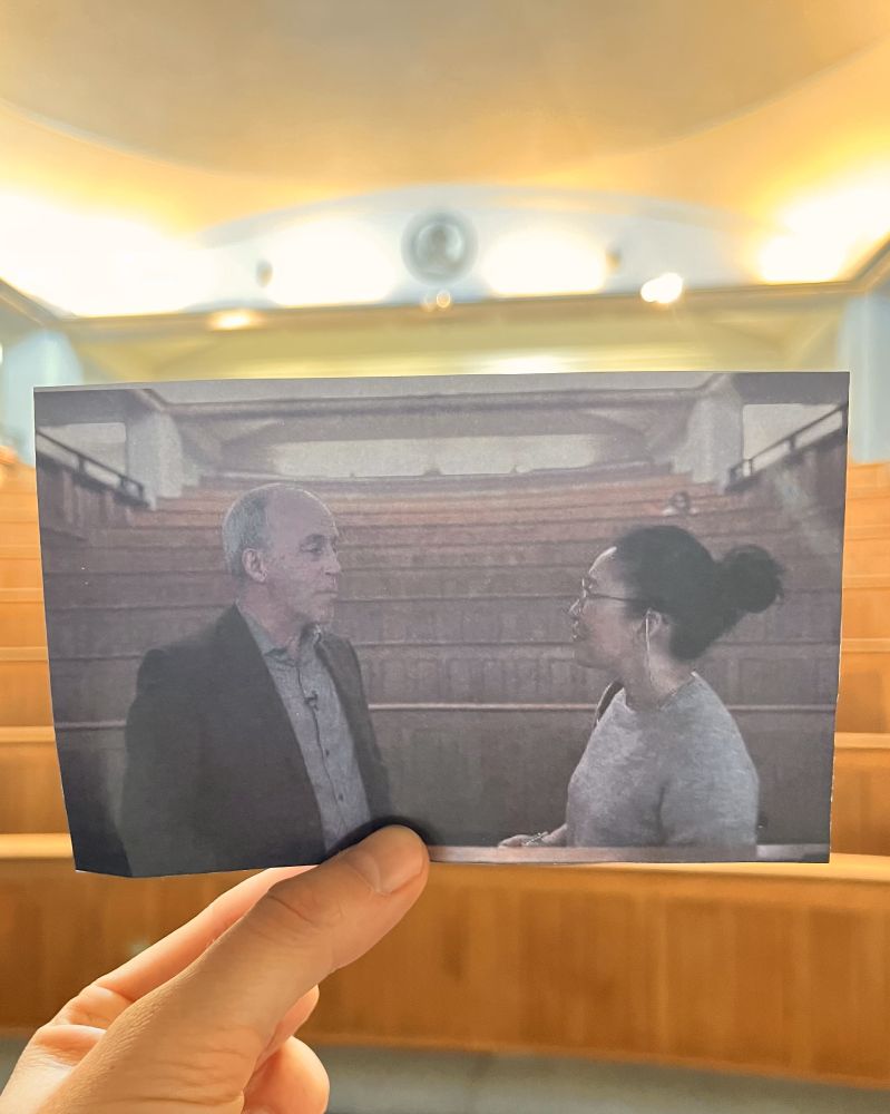 A person holds up a photograph of a scene from the movie "Killing Eve",  aligning it with the actual filming location visible in the background.
