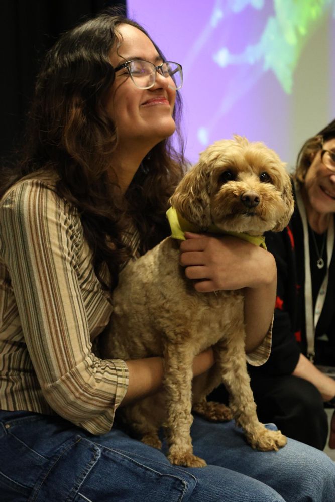 A person smiling and holding a small dog at a therapy session, with another attendee visible in the background.