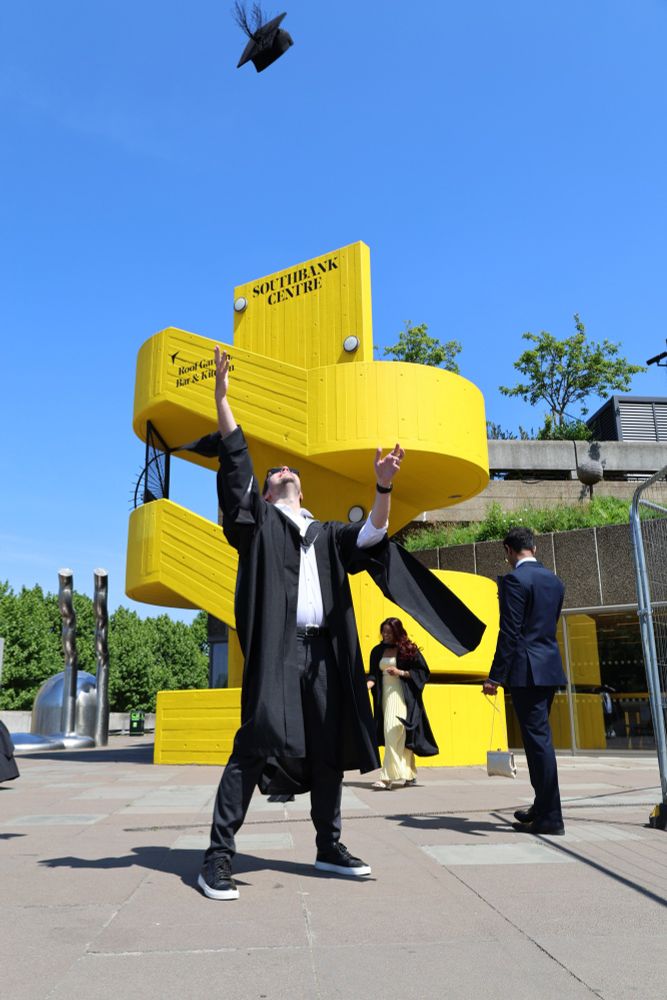 A person in a graduation gown joyfully tossing a mortarboard in the air in front of the yellow, sculptural sign of the Southbank Centre under a clear blue sky.