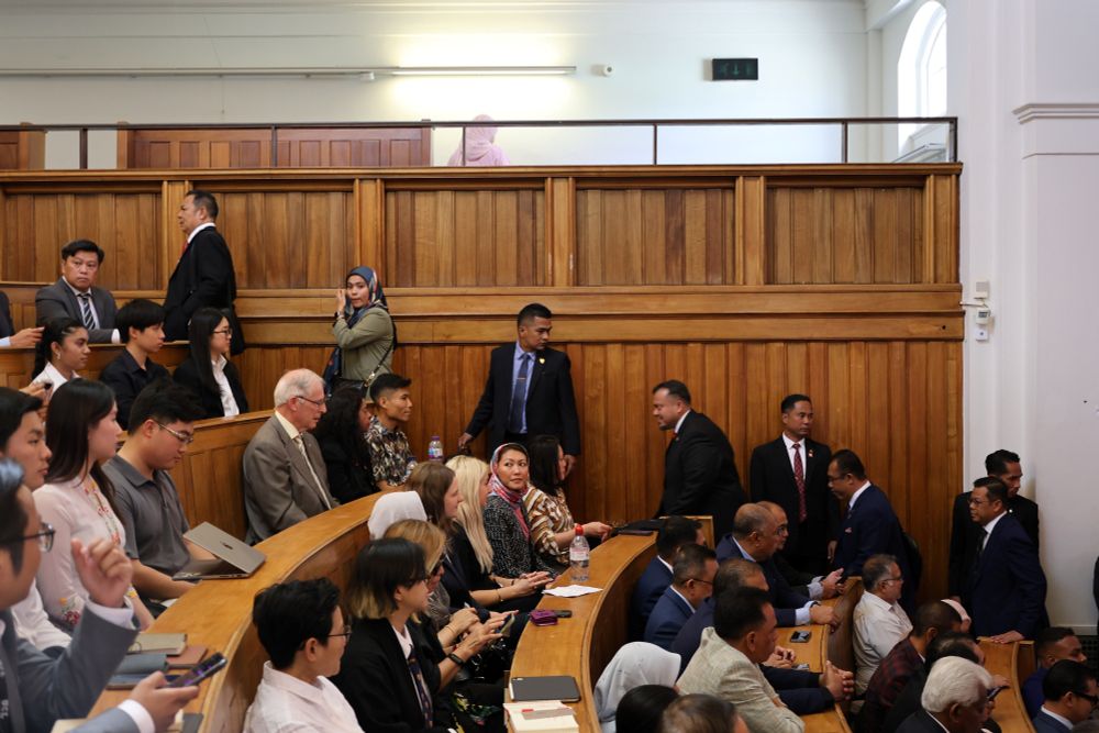Attendees in a crowded courtroom with wooden paneling, featuring various people in formal attire, including some individuals who are standing and others seated.