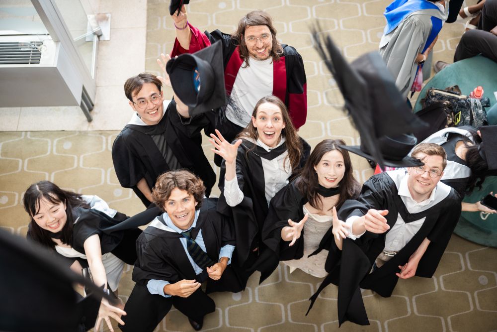 Group of smiling graduates in caps and gowns celebrating and throwing their caps in the air, looking up at the camera.