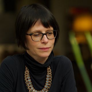 Kristin Bakke has short black hair and glasses wearing a black turtleneck and a decorative necklace, focusing thoughtfully in a softly lit environment.