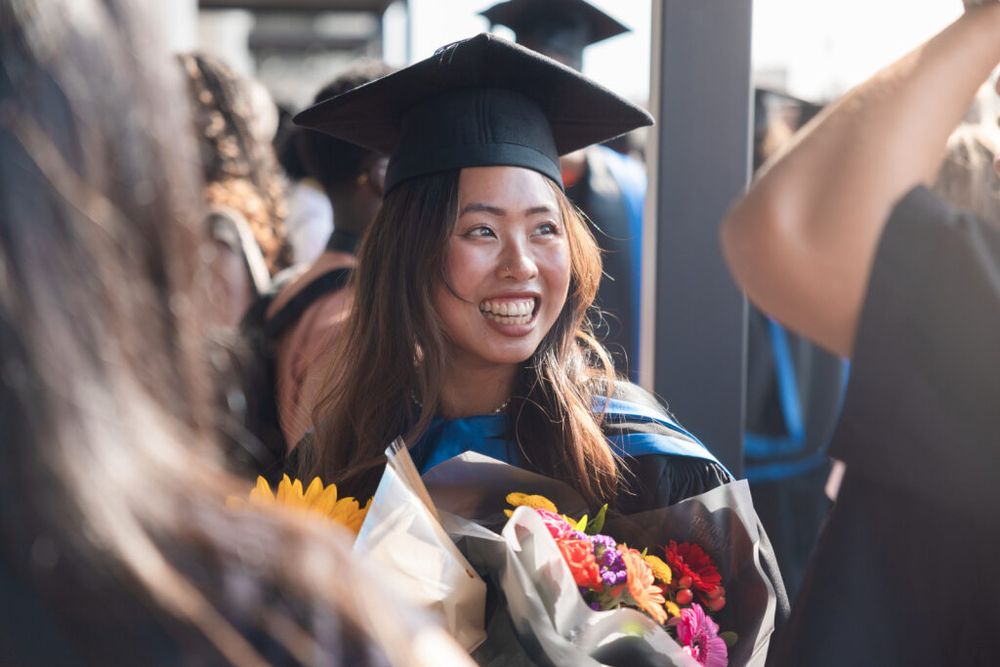 A joyful graduate in cap and gown, holding a bouquet of flowers, celebrates among peers.