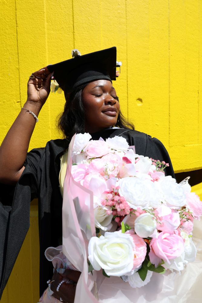 Graduate in academic regalia holding a large bouquet of pink and white flowers against a yellow background.