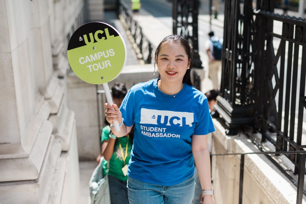 A UCL student ambassador holding a "UCL Campus Tour" sign and smiling, standing outside on a sunny day.