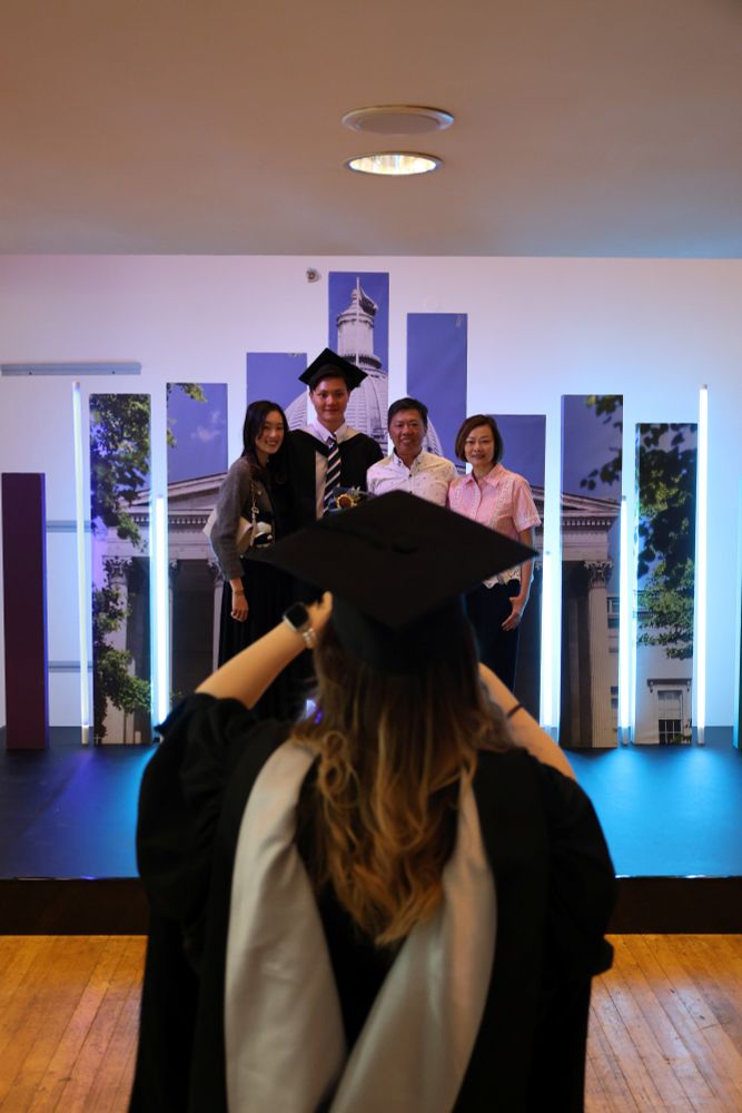 Graduate in cap and gown seen from behind, taking a photo of a group of three family members posing with another graduate in regalia, in a decorated room possibly at a university graduation ceremony.