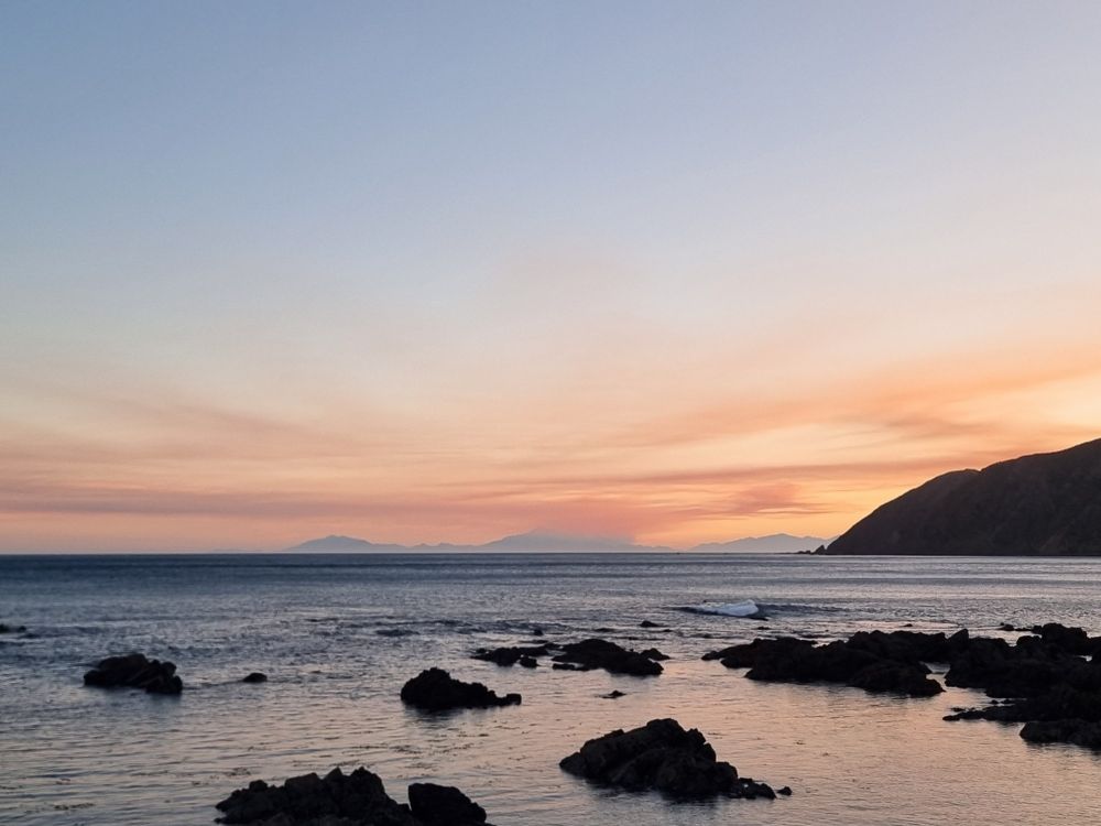 Immensely calm sea at the end of a day. The water is flat and reflective around isolated dark rocks in the foreground, the sky suffusing with orange-pink along the horizon, where three hazy distant mountains lurk. A closer, steep slope of hill is silhouette at right. 