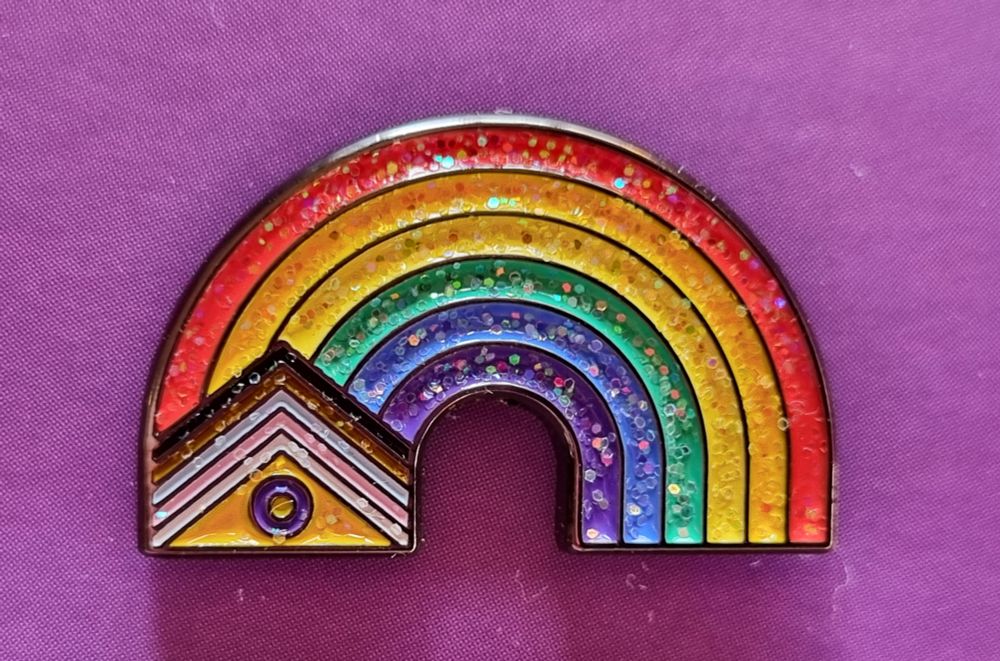 A 'Sweat With Pride' enamel pin, on a pink-purple background. It's a glittery rainbow arch, with overlapping triangles comprised of the rest of the colours of the fully inclusive Pride flag at one end (black, brown, pale blue, pale pink, white, and a yellow front triangle with a dark purple ring)