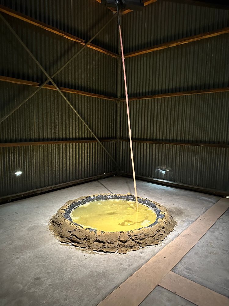 A long stick skating across a murky pool of a yellow substance, contained in a round brown receptacle. This exhibit is inside a large shed.