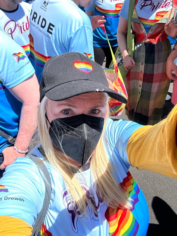 A person with long blonde hair wearing a rainbow shirt, a black cap with a rainbow heart, and a black facemask

