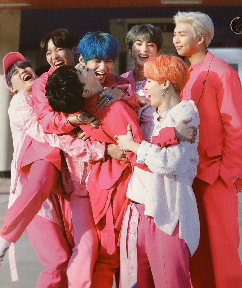 Pink dressed BTS members in bright hair colors do a group hug on the set of the "Boy with Love" MV