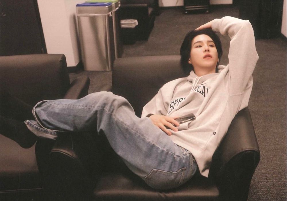 Yoongi in an off-white sweater and grey pants is lounging on brown leather armchair and pushed back his long black hair with his left hand