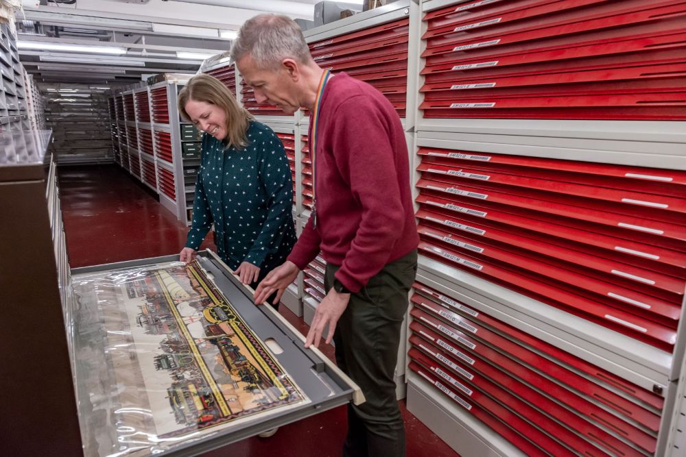 Two people in a repository opening a drawer and examining a large colourful illustration.