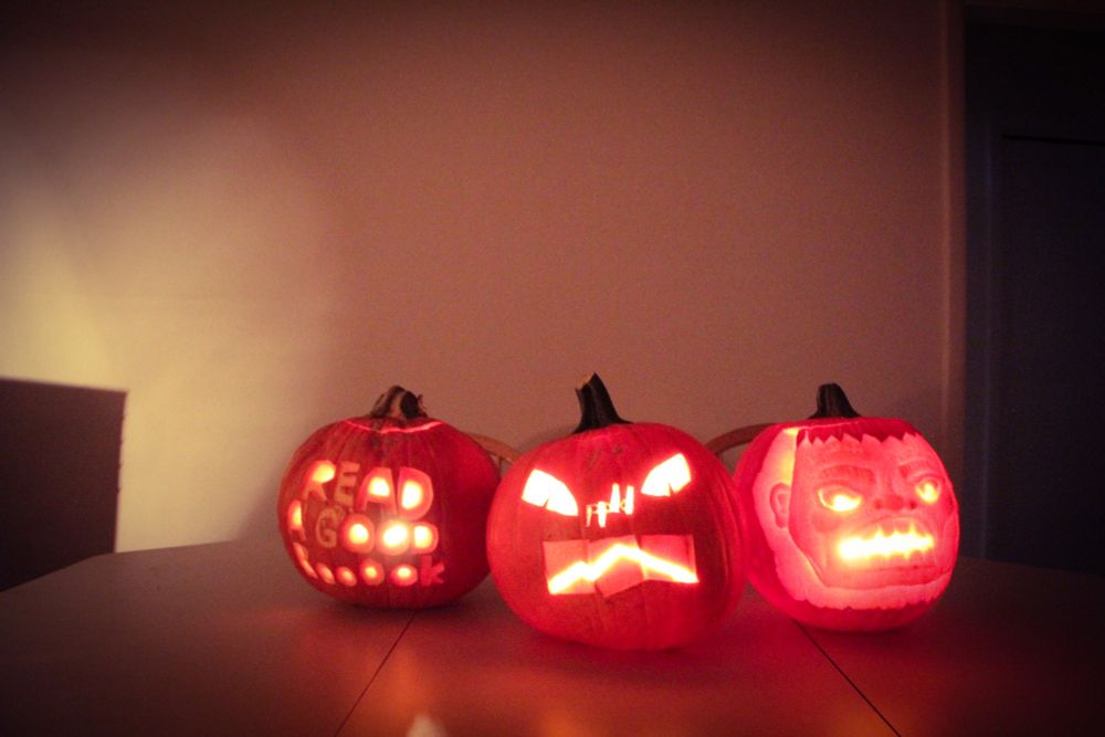 Three glowing jack-o-lanterns carved by a member of PPLD staff. One features the words "Read a Good Booook," one features the Library District's logo, and one features the face of Frankenstein's monster.