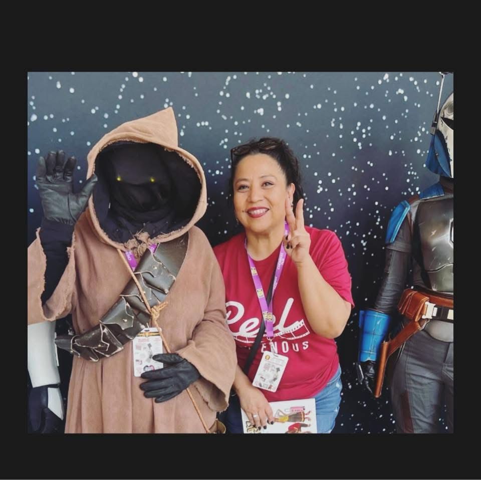 A creature in a brown hood stands next to a brown woman in a red shirt, holding up their fingers in a peace sign. 