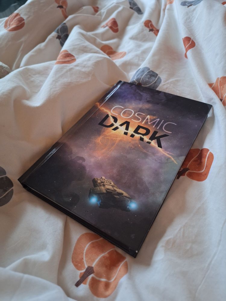 A copy of Cosmic Dark, a glossy book with a spaceship in the front, on a duvet with pumpkins on it.