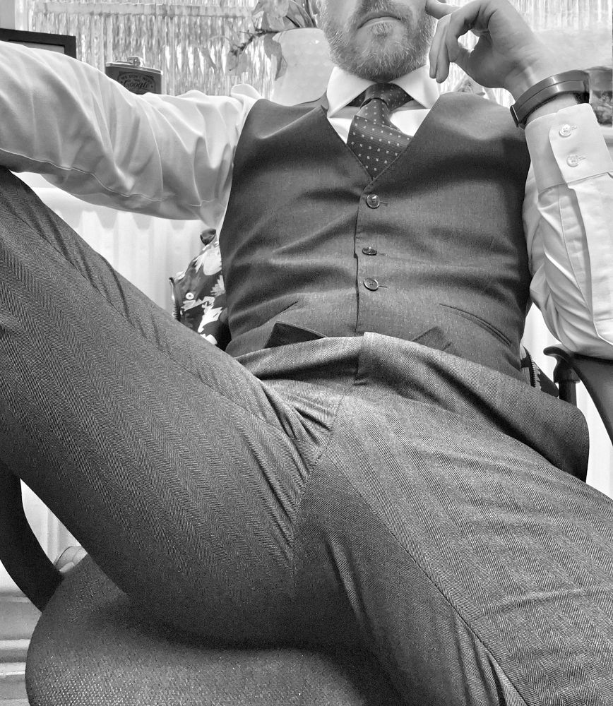 A black and white photo of a man dressed in a white shirt and tie, black waistcoat and trousers. His finger is on his temple and though only his mouth is visible in the frame, he looks moody.