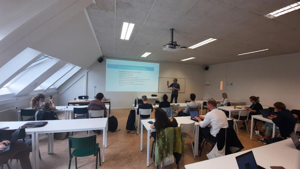 Image of a room with white walls and ceiling, people sitting in white desks and chairs and looking at Olivier Hoedeman as he does a powerpoint presentation.