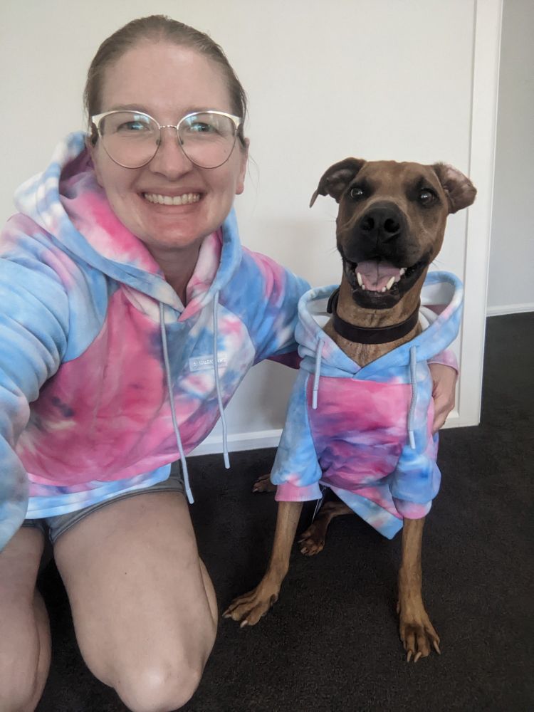 A white female wearing glasses and a tie die pink and blue hoodie kneels on the ground beside a ginger dog also wearing a pink and blue tie die hoodie. Both are smiling 