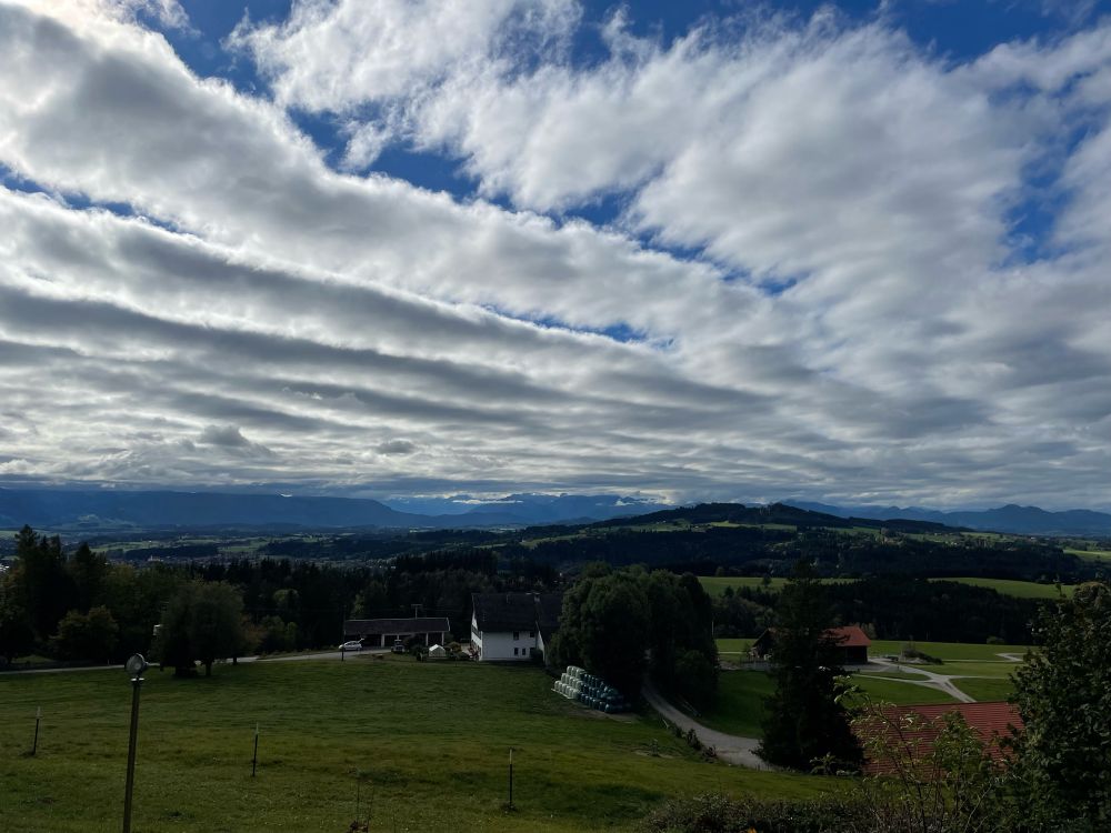 Blick vom Gipfel des Weichbergs zum Auerberg und in die Alpen. Es ist bewölkt, die Wolken links sehen aus, als wären sie mit einem Rechen in saubere Reihen arrangiert. Rechts zerfleddern sie.