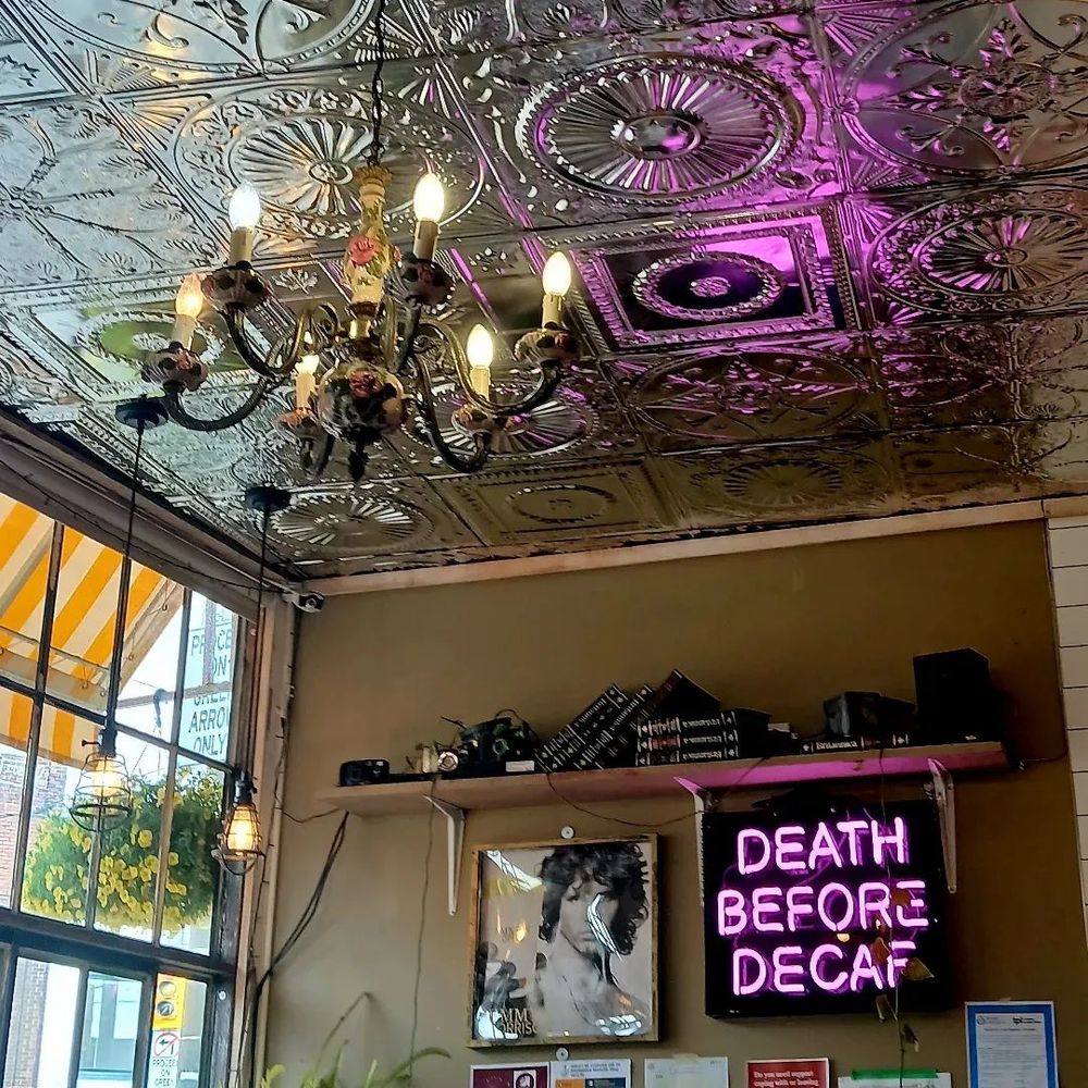 A pink neon sign mounted on a café wall says death before decaf. Beside it is a picture of Jim Morrison and above both these items there is a shelf with speakers, books and various other items. These all are mounted on a light brown wall above which there is a shiny tin ceiling with an elaborate chandelier. The café's front window is on the left;  outside there are a hanging basket of yellow flowers and a yellow and white striped awning. 