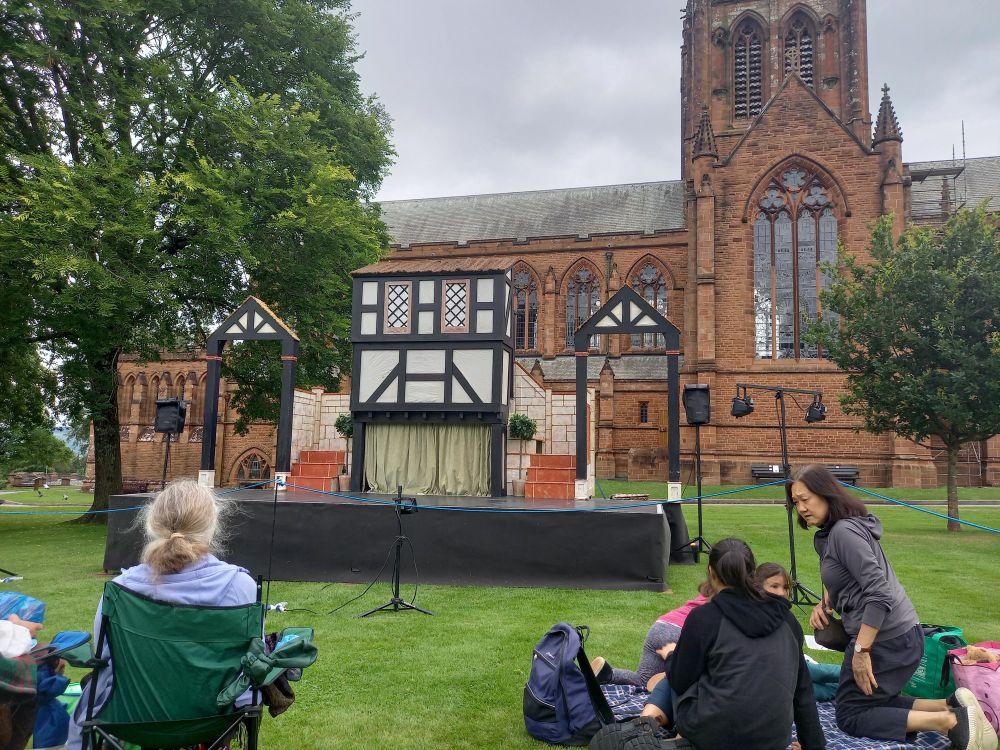 An outdoor scene with a stage with a Tudor house front.  Behind a large sandstone church.