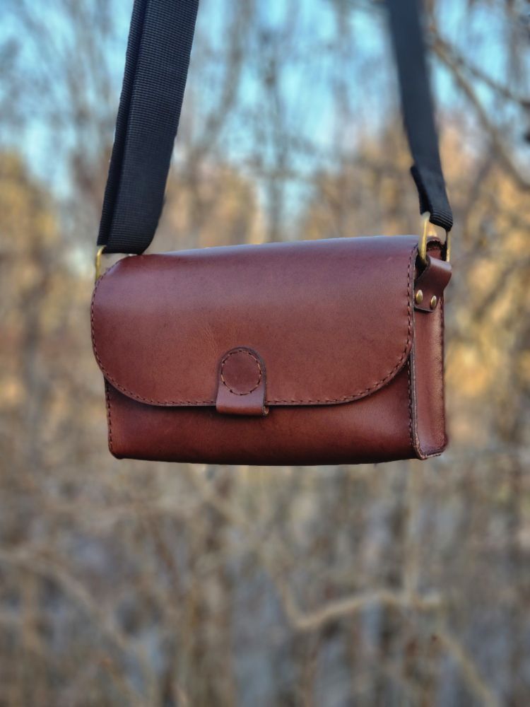 A chocolate brown bag are in focus, hanging in front of a hedge without leaves. Its rectangular and broader than its high, the size is just big enough to keep a mobile phone and a wallet in it. the string is black and looks like a car belt just a bit more narrow. In the background, thru the hedge, you can see some blur trees and a blue sky. 