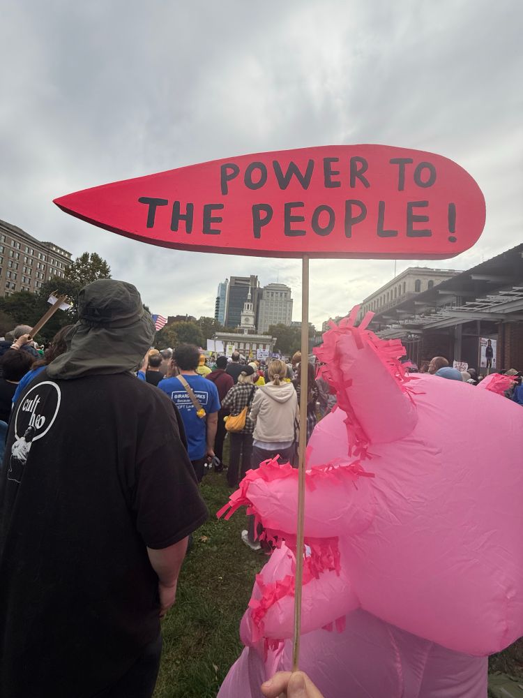 A picture of the Philadelphia no kings protest on the mall in front of Independence Hall. It’s a crowd shot with the Hall in the distance. In the foreground is a pink Pokémon looking thing and I’m holding up a pink speech bubble that says “power to the people at the top of the picture. 
