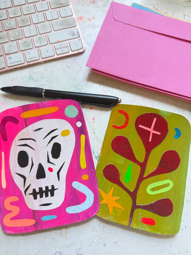 A picture of two cards, a black pen, a pile of pink envelopes set near the corner of a keyboard. The card on the bottom left is pink with a white skull surrounded by colorful random shapes. The card next to it on the right is green with an abstract maroon plant also with abstract shapes surrounding it. Art is made with painted cut paper by artist Martha Rich. 