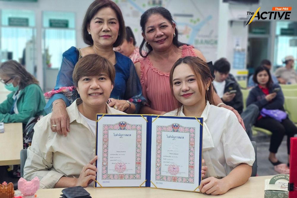 Ae and ying both wear light colored shirts and hold up their wedding license. Behind them are their mothers.