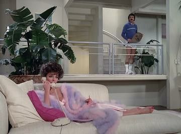 A woman laying on the couch in a purple & pink robe while talking on the landline.