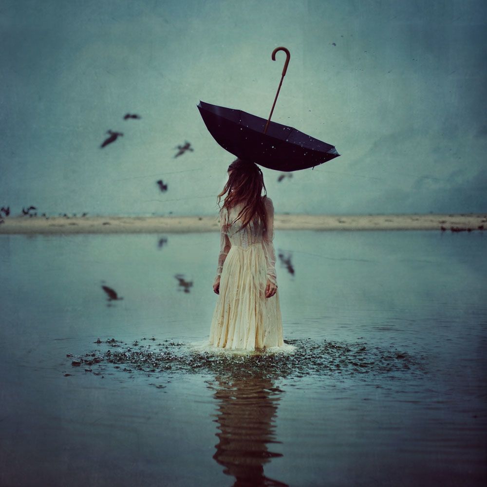 A woman in a white dress standing in a lake with an upside down umbrella on her head and blurry black birds flying around 