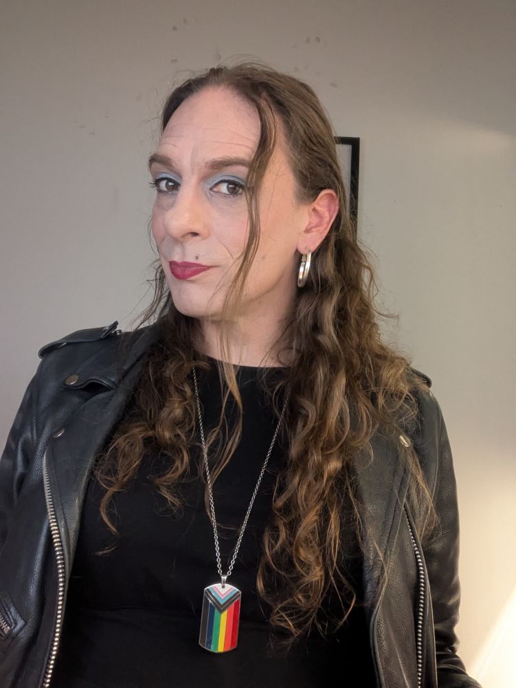 Jessie looking pretty great (Even if she does alt text so herself), all in black with a biker jacket and a progress pride pendant. Hair down, not bad make up and a strong red lip.