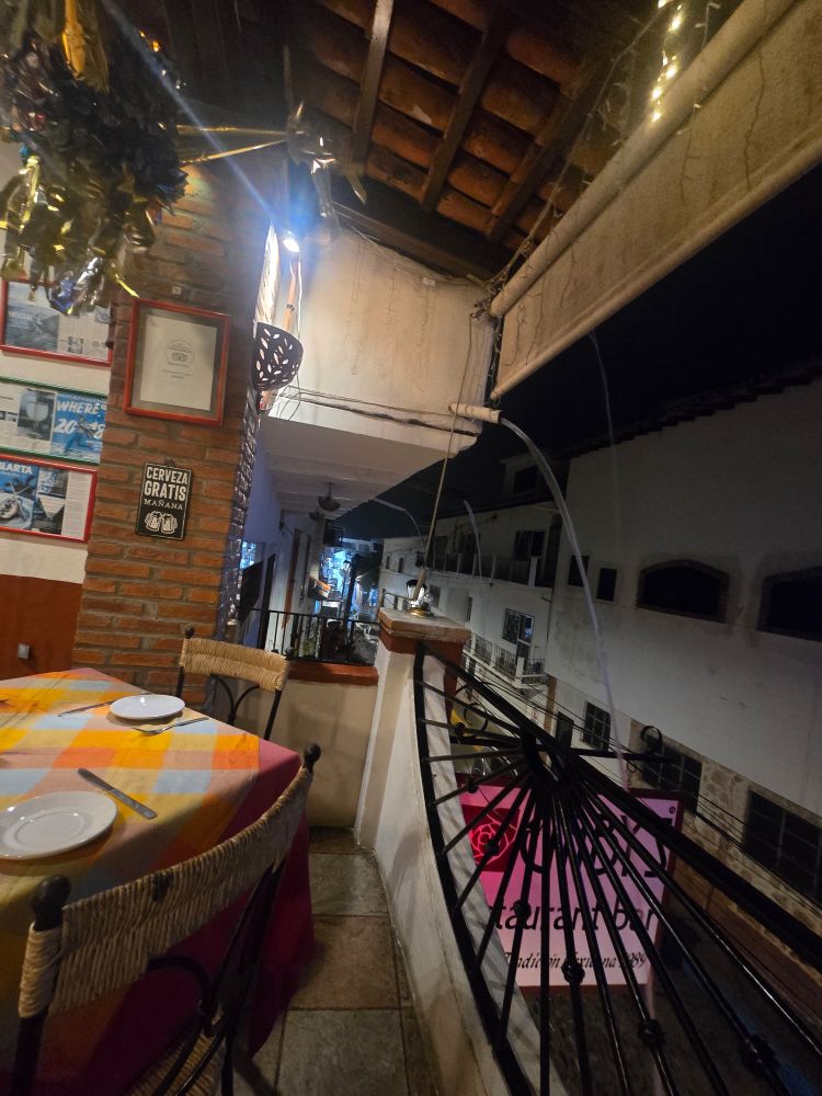 A restaurant awning on the 2nd floor with tables and chairs with bright tablecloths with rainwater gutters pouring water in small fountains into the streets