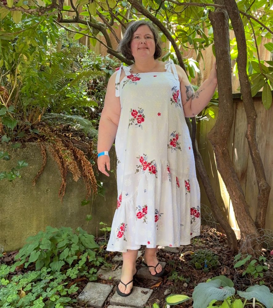 A woman in a maxi length white sundress with red flowers, facing the camera straight on and standing beside a tree