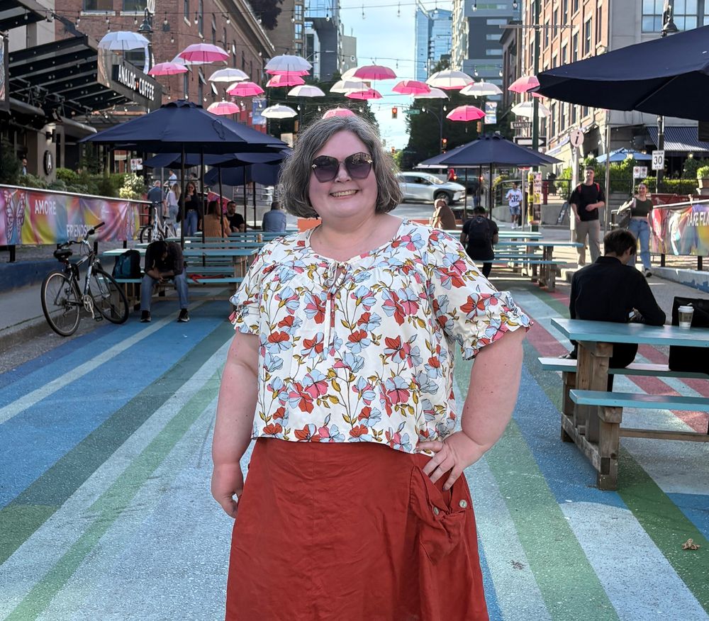 A woman in a short sleeved loose blouse in a floral print fabric standing on a city street with umbrellas hanging overhead, stripes painted on the ground, and picnic tables with people sitting at them 