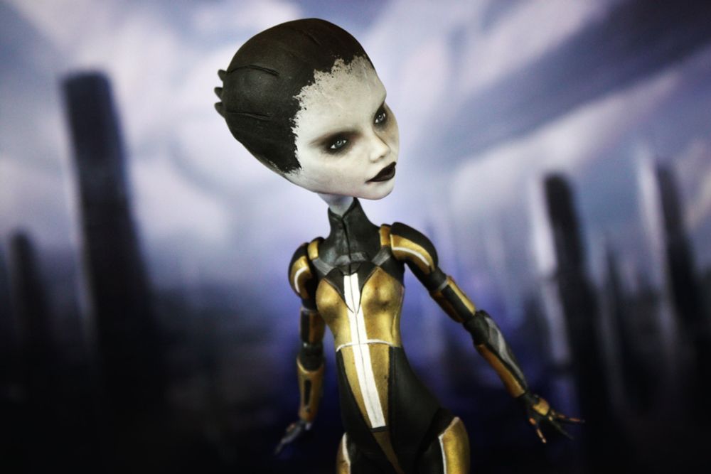 Una foto que hice a la muñeca Monster High customizada/modificada/pintada por mi de la que hablo en el post. Se trata de una Asari de piel gris con un uniforme tono dorado y negro con una franja blanca, su clase era la de Adepto. Posa frente a una imagen de la Ciudadela del juego Mass Effect, que es una ciudad de edificios altos y futuristas.