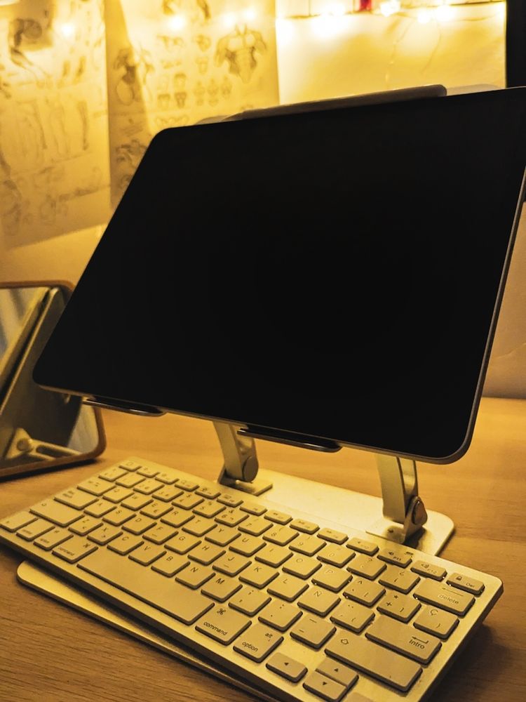 Una foto rápida a mi escritorio con el soporte de metal y el iPad colocado que comento a Sabro en el post. Bajo el iPad está el teclado. Se ve el resto del escritorio, que es de madera y tiene un espejo a un lado. Al fondo se ve luces amarillas y apuntes de anatomía que uso para trabajar.