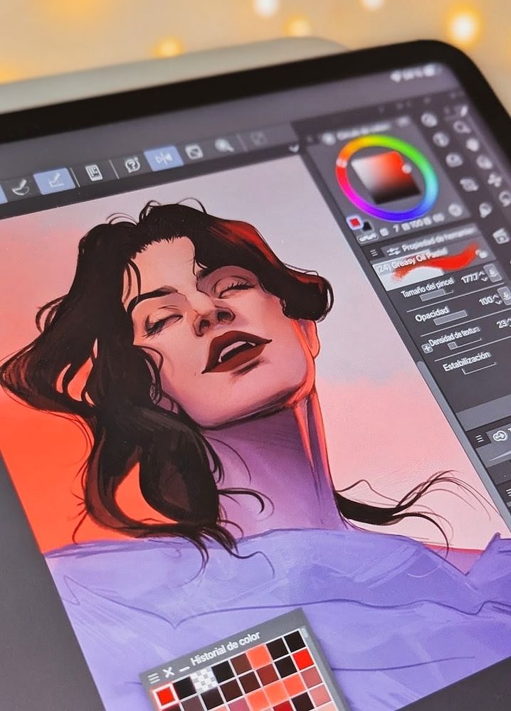 Foto a la pantalla de mi iPad con un dibujo en proceso de mi personaje Sagrado, de mi cómic Coyote Tresvidas, una vampira de pelo oscuro y en el dibujo ojos blancos enfocada desde abajo. El dibujo tiene una luz roja anaranjada g distintos tonos azules. El programa que se está usando es Clip studio paint, aparece en el menú de herramienta uno de los pinceles de los que hablo. De fondo, detrás de la pantalla, unas luces amarillas de mi escritorio.