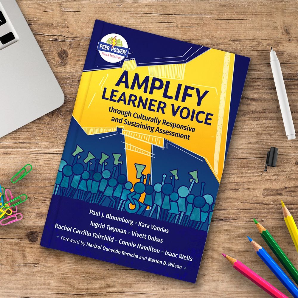 The book Amplify Learner Voice through Culturally Responsive and Sustaining Assessment on a wooden table with the edge of a computer, some colored pencils, colorful paperclips and a white pen around it