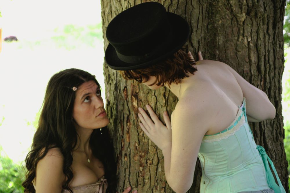 me as rosalind in a pink corset and rose hairpins, staring up at maijai as thalia in a matching green corset and top hat from around the trunk of a green amidst bright greenery